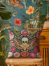 A square decorative throw pillow with a dark navy background and a linen-feel polyester texture. The design features a large, centrally-placed Death's Head hawkmoth with a detailed human skull marking on its thorax. The moth's wings are intricately patterned in shades of red, green, and gold. It is surrounded by a dense, symmetrical arrangement of colorful folk-art style flowers in vibrant shades of pink, red, and orange, with green leaves and vines. The print has a faux-embroidery appearance.