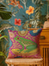 A square decorative throw pillow made from premium polyester with a visible linen-like woven texture. The background is a vibrant hot pink covered in a dense, retro-style floral pattern in orange, teal, and gold. The main design is a brightly colored, stylized illustration of a green red-eyed tree frog with orange feet and prominent red eyes. The whimsical frog is riding a vibrant yellow banana. The overall aesthetic is colorful, maximalist, and eclectic, with a faux-embroidered texture effect.