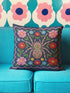 Square throw pillow cover featuring a symmetrical folk art spider illustration centered on a large hot pink flower. The spider has a striped abdomen in green, red, and yellow with pink legs. The background is a textured dark navy blue fabric with a visible faux linen weave. Surrounding the central spider are stylized botanical motifs including red daisies, blue forget-me-nots, and green leaves. The illustration is flat printed with a high-quality finish. This image shows the case only.