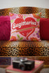 A square decorative throw pillow featuring a light pink background with a trompe-l'œil faux-embroidery print. A large red heart sits in the center, pierced by a green and white arrow. The word Sagittarius is printed across the heart in a puffy white retro font with a red drop shadow. The central design is surrounded by scattered vintage-style flowers in vibrant shades of purple, orange, and yellow with bright green leaves. The texture is a flat, high-fidelity printed illusion.