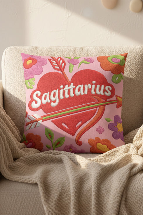 A square decorative throw pillow featuring a light pink background with a trompe-l'œil faux-embroidery print. A large red heart sits in the center, pierced by a green and white arrow. The word Sagittarius is printed across the heart in a puffy white retro font with a red drop shadow. The central design is surrounded by scattered vintage-style flowers in vibrant shades of purple, orange, and yellow with bright green leaves. The texture is a flat, high-fidelity printed illusion.