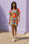 Short sleeve crew neck t-shirt dress on a cream background with an allover retro mod flower medallion print. Bold 60s daisies in red, yellow, and white sit at the center of concentric rainbow rings in lime green, hot pink, orange, and royal blue, with small magenta squares and tiny red checkerboard accents between the motifs. Above-the-knee straight fit silhouette in a 95 percent cotton, 5 percent elastane heavyweight stretch blend. Groovy retro flower power dress for dopamine dressing and easy summer outfi