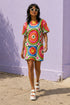Short sleeve crew neck t-shirt dress on a cream background with an allover retro mod flower medallion print. Bold 60s daisies in red, yellow, and white sit at the center of concentric rainbow rings in lime green, hot pink, orange, and royal blue, with small magenta squares and tiny red checkerboard accents between the motifs. Above-the-knee straight fit silhouette in a 95 percent cotton, 5 percent elastane heavyweight stretch blend. Groovy retro flower power dress for dopamine dressing and easy summer outfi