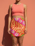 Form-fitting mini skirt featuring a vibrant, retro-inspired maximalist pattern. The design showcases a diagonal hot pink band filled with white and yellow daisies, contrasting against a bold background of wavy, abstract red and yellow flame-like stripes. The bottom edge includes curved color blocks of green and orange. Made of a smooth, stretchy fabric with a mid-rise elastic waistband, hitting at mid-thigh. Available in both a matte black-light reactive finish and a metallic glitter finish.