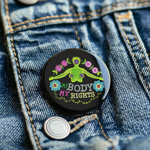 Round black pinback button featuring lime green female figure with outstretched arms and purple floral details, surrounded by pink flowers and blue blooms with multicolored "MY BODY MY RIGHTS" text. Folk art feminist pin with glossy finish and safety pin backing in 2" and 3" sizes.