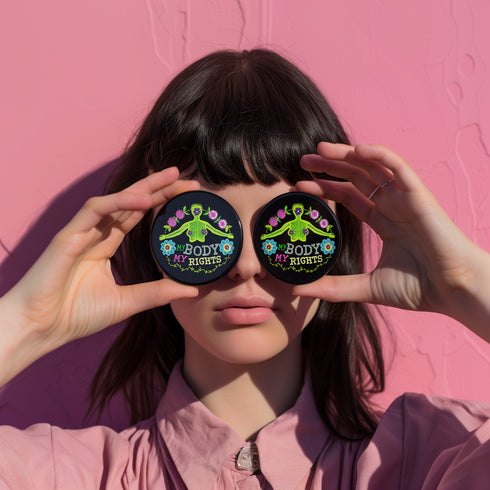 Round black pinback button featuring lime green female figure with outstretched arms and purple floral details, surrounded by pink flowers and blue blooms with multicolored "MY BODY MY RIGHTS" text. Folk art feminist pin with glossy finish and safety pin backing in 2" and 3" sizes.