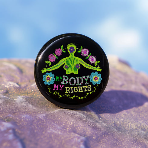 Round black pinback button featuring lime green female figure with outstretched arms and purple floral details, surrounded by pink flowers and blue blooms with multicolored "MY BODY MY RIGHTS" text. Folk art feminist pin with glossy finish and safety pin backing in 2" and 3" sizes.