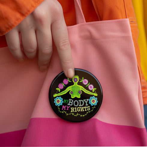 Round black pinback button featuring lime green female figure with outstretched arms and purple floral details, surrounded by pink flowers and blue blooms with multicolored "MY BODY MY RIGHTS" text. Folk art feminist pin with glossy finish and safety pin backing in 2" and 3" sizes.