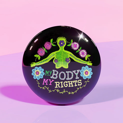 Round black pinback button featuring lime green female figure with outstretched arms and purple floral details, surrounded by pink flowers and blue blooms with multicolored "MY BODY MY RIGHTS" text. Folk art feminist pin with glossy finish and safety pin backing in 2" and 3" sizes.