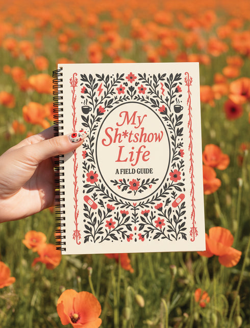 An 8.5 x 11 inch hardcover spiral-bound planner with a cream-colored cover featuring red and black folk art style floral illustrations. The cover reads "My Sh*tshow Life: A Field Guide" in a retro serif font. The planner has a durable satin matte laminated cover adhered to chipboard and a metal wire-o spiral binding available in gold or black. Interior pages are undated and include monthly and weekly layouts with sections for notes, to-do lists.