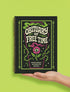 A black hardcover spiral planner. The cover features a vibrant, coffin-shaped frame in lime green and magenta pink, bordered by thorny vines. Inside the frame, the title "Obituary of Free Time" is written in a psychedelic 70s-style font. Below the title, a pink flower with a single green eyeball at its center blooms from a thorny stem. The planner is bound with a gold metal wire-o spiral on the left side. The design has a spooky cute, gothic floral aesthetic.