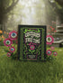 A black hardcover spiral planner. The cover features a vibrant, coffin-shaped frame in lime green and magenta pink, bordered by thorny vines. Inside the frame, the title "Obituary of Free Time" is written in a psychedelic 70s-style font. Below the title, a pink flower with a single green eyeball at its center blooms from a thorny stem. The planner is bound with a gold metal wire-o spiral on the left side. The design has a spooky cute, gothic floral aesthetic.