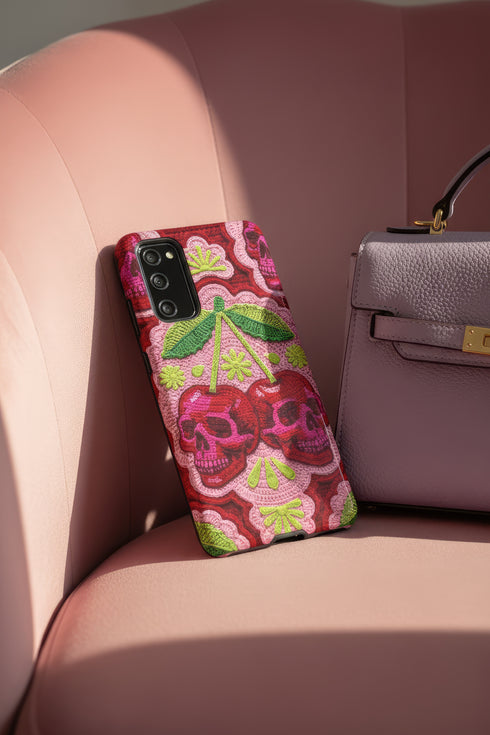 Dual layer matte phone case with a high fidelity 2D print that creates a hyper realistic faux crochet and knit illusion. The central design shows two conjoined cherries styled as bright pink and burgundy human skulls with lime green stems and leaves. The background is a detailed scalloped lace doily pattern in hot pink, deep red, and lime green. Matte finish shell with a black inner TPU lining, precise camera cutout, and open ports. Pastel goth macabre alt fashion phone accessory, made to order.