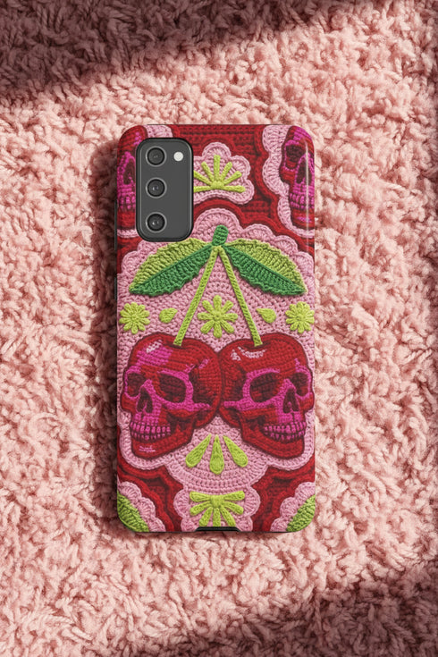 Dual layer matte phone case with a high fidelity 2D print that creates a hyper realistic faux crochet and knit illusion. The central design shows two conjoined cherries styled as bright pink and burgundy human skulls with lime green stems and leaves. The background is a detailed scalloped lace doily pattern in hot pink, deep red, and lime green. Matte finish shell with a black inner TPU lining, precise camera cutout, and open ports. Pastel goth macabre alt fashion phone accessory, made to order.