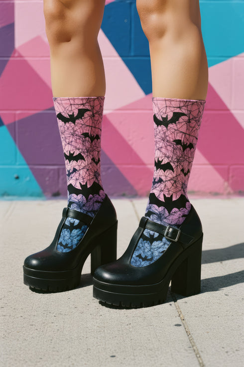 A pair of crew length socks featuring a vibrant pastel gradient background that transitions from light pink at the top, to purple in the middle, and light blue at the toe. Overlaid on this gradient is a solid black, intricate spiderweb pattern that covers the entire sock. Scattered throughout the spiderweb design are multiple black silhouettes of flying bats in various sizes. The socks have a solid black reinforced heel and toe, made of a smooth, stretchy, tight knit fabric.