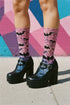 A pair of crew length socks featuring a vibrant pastel gradient background that transitions from light pink at the top, to purple in the middle, and light blue at the toe. Overlaid on this gradient is a solid black, intricate spiderweb pattern that covers the entire sock. Scattered throughout the spiderweb design are multiple black silhouettes of flying bats in various sizes. The socks have a solid black reinforced heel and toe, made of a smooth, stretchy, tight knit fabric.