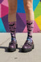 A pair of crew length socks featuring a vibrant pastel gradient background that transitions from light pink at the top, to purple in the middle, and light blue at the toe. Overlaid on this gradient is a solid black, intricate spiderweb pattern that covers the entire sock. Scattered throughout the spiderweb design are multiple black silhouettes of flying bats in various sizes. The socks have a solid black reinforced heel and toe, made of a smooth, stretchy, tight knit fabric.