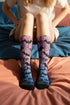 A pair of crew length socks featuring a vibrant pastel gradient background that transitions from light pink at the top, to purple in the middle, and light blue at the toe. Overlaid on this gradient is a solid black, intricate spiderweb pattern that covers the entire sock. Scattered throughout the spiderweb design are multiple black silhouettes of flying bats in various sizes. The socks have a solid black reinforced heel and toe, made of a smooth, stretchy, tight knit fabric.