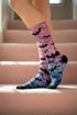 A pair of crew length socks featuring a vibrant pastel gradient background that transitions from light pink at the top, to purple in the middle, and light blue at the toe. Overlaid on this gradient is a solid black, intricate spiderweb pattern that covers the entire sock. Scattered throughout the spiderweb design are multiple black silhouettes of flying bats in various sizes. The socks have a solid black reinforced heel and toe, made of a smooth, stretchy, tight knit fabric.