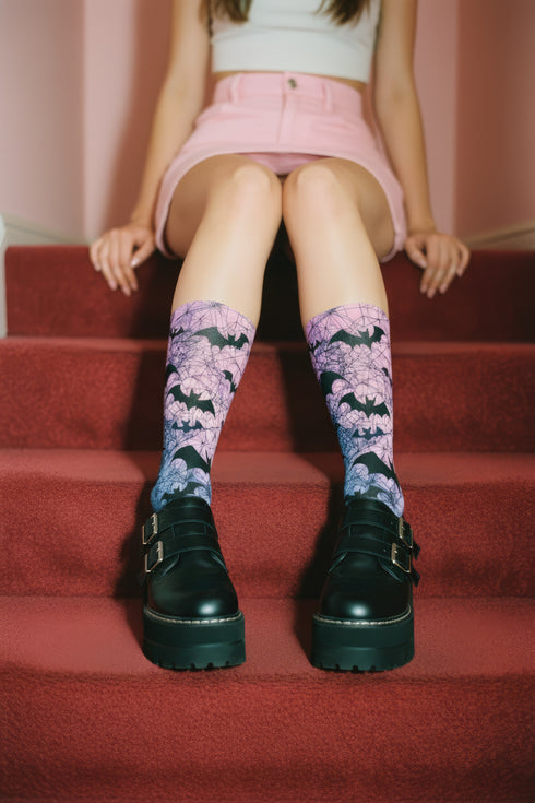 A pair of crew length socks featuring a vibrant pastel gradient background that transitions from light pink at the top, to purple in the middle, and light blue at the toe. Overlaid on this gradient is a solid black, intricate spiderweb pattern that covers the entire sock. Scattered throughout the spiderweb design are multiple black silhouettes of flying bats in various sizes. The socks have a solid black reinforced heel and toe, made of a smooth, stretchy, tight knit fabric.