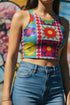A high-resolution product photo of a body-hugging, 2D printed crop top with a 1970s-inspired floral patchwork pattern. The design consists of vibrant squares in cobalt blue, pale lavender, and deep violet, separated by pink borders with red dots. Central motifs include yellow daisies with orange centers and green-magenta floral mandalas. The top is made of a smooth, high-stretch performance knit fabric with curved back armholes and a cropped waistline.
