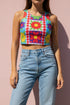 A high-resolution product photo of a body-hugging, 2D printed crop top with a 1970s-inspired floral patchwork pattern. The design consists of vibrant squares in cobalt blue, pale lavender, and deep violet, separated by pink borders with red dots. Central motifs include yellow daisies with orange centers and green-magenta floral mandalas. The top is made of a smooth, high-stretch performance knit fabric with curved back armholes and a cropped waistline.