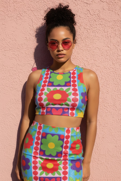 A high-resolution product photo of a body-hugging, 2D printed crop top with a 1970s-inspired floral patchwork pattern. The design consists of vibrant squares in cobalt blue, pale lavender, and deep violet, separated by pink borders with red dots. Central motifs include yellow daisies with orange centers and green-magenta floral mandalas. The top is made of a smooth, high-stretch performance knit fabric with curved back armholes and a cropped waistline.