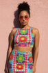 A high-resolution product photo of a body-hugging, 2D printed crop top with a 1970s-inspired floral patchwork pattern. The design consists of vibrant squares in cobalt blue, pale lavender, and deep violet, separated by pink borders with red dots. Central motifs include yellow daisies with orange centers and green-magenta floral mandalas. The top is made of a smooth, high-stretch performance knit fabric with curved back armholes and a cropped waistline.