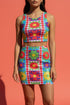 A high-resolution product photo of a body-hugging, 2D printed crop top with a 1970s-inspired floral patchwork pattern. The design consists of vibrant squares in cobalt blue, pale lavender, and deep violet, separated by pink borders with red dots. Central motifs include yellow daisies with orange centers and green-magenta floral mandalas. The top is made of a smooth, high-stretch performance knit fabric with curved back armholes and a cropped waistline.