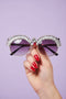 Sunglasses with decorative metal frame held by a hand against a purple background