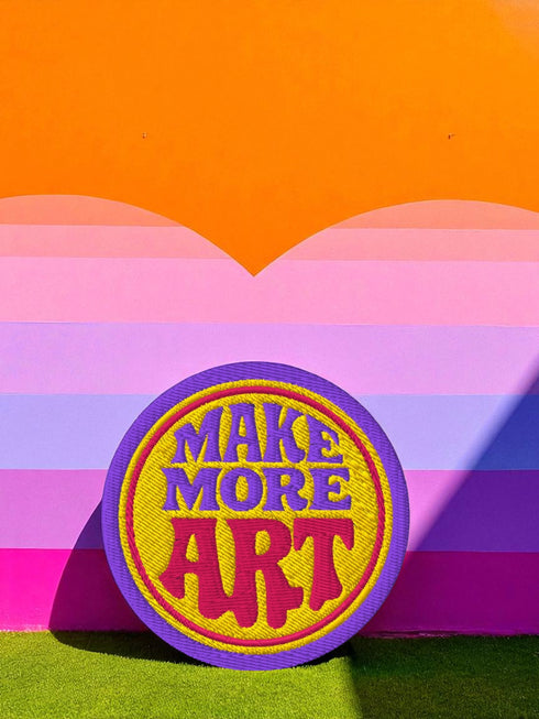 A round embroidered patch with the words "MAKE MORE ART" in a retro 70s psychedelic font. The word "ART" is magenta pink, "MAKE MORE" is purple, and they are set against a bright yellow circular background. The entire design is encircled by a thin pink line and a thick purple border. This 3-inch iron-on applique is a colorful, groovy accessory for DIY projects, perfect for personalizing jackets, backpacks, or denim vests. It serves as an inspirational artist gift with a vintage aesthetic.