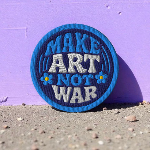 Round embroidered iron-on patch featuring "MAKE ART NOT WAR" text with blue floral design. Navy blue background with light blue border, white cream lettering, and small yellow-centered blue flowers. 3-inch circular twill fabric patch perfect for jackets, backpacks, denim, and clothing customization. Anti-war peace activist feminist punk alternative accessory for creative counterculture gift giving.