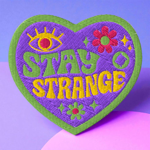 A detailed embroidered patch in the shape of a purple heart with a vibrant green border. In a groovy, psychedelic font, the words "STAY STRANGE" are stitched in yellow and green. The design includes a mystical third eye at the top, and small retro flowers and stars, giving it a 70s hippie vibe.