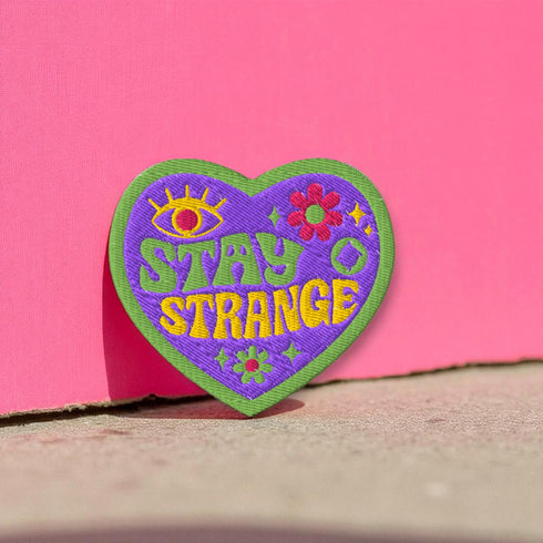 A detailed embroidered patch in the shape of a purple heart with a vibrant green border. In a groovy, psychedelic font, the words "STAY STRANGE" are stitched in yellow and green. The design includes a mystical third eye at the top, and small retro flowers and stars, giving it a 70s hippie vibe.