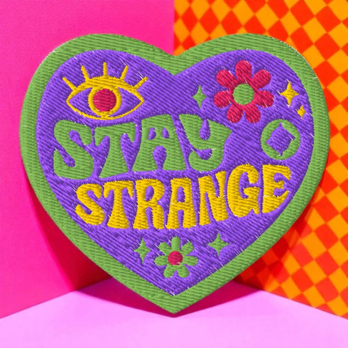 A detailed embroidered patch in the shape of a purple heart with a vibrant green border. In a groovy, psychedelic font, the words "STAY STRANGE" are stitched in yellow and green. The design includes a mystical third eye at the top, and small retro flowers and stars, giving it a 70s hippie vibe.