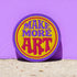 A round embroidered patch with the words "MAKE MORE ART" in a retro 70s psychedelic font. The word "ART" is magenta pink, "MAKE MORE" is purple, and they are set against a bright yellow circular background. The entire design is encircled by a thin pink line and a thick purple border. This 3-inch iron-on applique is a colorful, groovy accessory for DIY projects, perfect for personalizing jackets, backpacks, or denim vests. It serves as an inspirational artist gift with a vintage aesthetic.