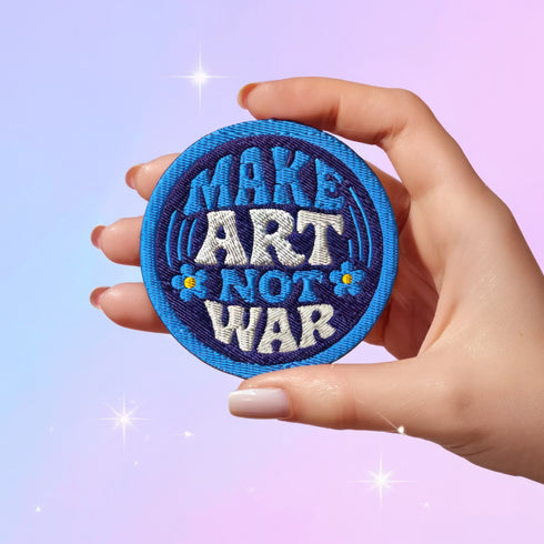 Round embroidered iron-on patch featuring "MAKE ART NOT WAR" text with blue floral design. Navy blue background with light blue border, white cream lettering, and small yellow-centered blue flowers. 3-inch circular twill fabric patch perfect for jackets, backpacks, denim, and clothing customization. Anti-war peace activist feminist punk alternative accessory for creative counterculture gift giving.