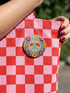 A circular folk art style 2.25 inch metal pinback button with a high-gloss finish. The design features a "NO WAR" text in magenta pink over a vibrant primrose yellow center with a sky blue peace sign motif. The design uses a "Trompe-l'œil" printed illusion to mimic embroidery and crochet stitching. The central medallion is surrounded by a scalloped pink ring and an outer ring of sunflowers. The back of the button is a durable silver metal tinplate with a secure safety pin fastening mechanism.