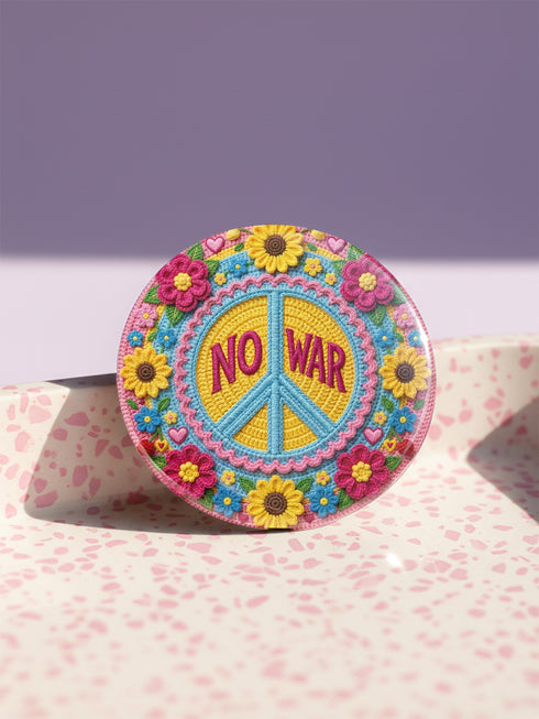 A circular folk art style 2.25 inch metal pinback button with a high-gloss finish. The design features a "NO WAR" text in magenta pink over a vibrant primrose yellow center with a sky blue peace sign motif. The design uses a "Trompe-l'œil" printed illusion to mimic embroidery and crochet stitching. The central medallion is surrounded by a scalloped pink ring and an outer ring of sunflowers. The back of the button is a durable silver metal tinplate with a secure safety pin fastening mechanism.