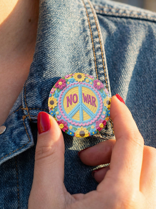 A circular folk art style 2.25 inch metal pinback button with a high-gloss finish. The design features a "NO WAR" text in magenta pink over a vibrant primrose yellow center with a sky blue peace sign motif. The design uses a "Trompe-l'œil" printed illusion to mimic embroidery and crochet stitching. The central medallion is surrounded by a scalloped pink ring and an outer ring of sunflowers. The back of the button is a durable silver metal tinplate with a secure safety pin fastening mechanism.