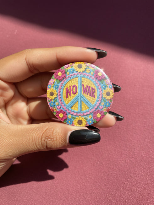 A circular folk art style 2.25 inch metal pinback button with a high-gloss finish. The design features a "NO WAR" text in magenta pink over a vibrant primrose yellow center with a sky blue peace sign motif. The design uses a "Trompe-l'œil" printed illusion to mimic embroidery and crochet stitching. The central medallion is surrounded by a scalloped pink ring and an outer ring of sunflowers. The back of the button is a durable silver metal tinplate with a secure safety pin fastening mechanism.