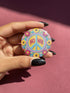 A circular folk art style 2.25 inch metal pinback button with a high-gloss finish. The design features a "NO WAR" text in magenta pink over a vibrant primrose yellow center with a sky blue peace sign motif. The design uses a "Trompe-l'œil" printed illusion to mimic embroidery and crochet stitching. The central medallion is surrounded by a scalloped pink ring and an outer ring of sunflowers. The back of the button is a durable silver metal tinplate with a secure safety pin fastening mechanism.
