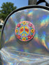A circular folk art style 2.25 inch metal pinback button with a high-gloss finish. The design features a "NO WAR" text in magenta pink over a vibrant primrose yellow center with a sky blue peace sign motif. The design uses a "Trompe-l'œil" printed illusion to mimic embroidery and crochet stitching. The central medallion is surrounded by a scalloped pink ring and an outer ring of sunflowers. The back of the button is a durable silver metal tinplate with a secure safety pin fastening mechanism.