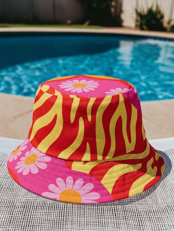 A reversible bucket hat with two distinct sides. Side one features a red and yellow zebra pattern with large pink and orange daisies. Side two has a deep purple background with white stars, rainbow scalloped bands in blue, green, and orange, and retro black and white flowers. The fabric has a visible textured linen weave. The hat is shown in a structured, professional studio shot against a white background, highlighting its 100% polyester durability.