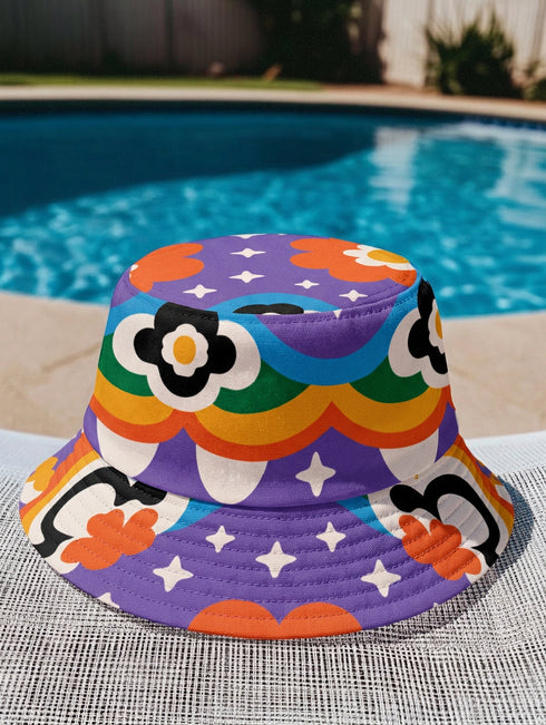 A reversible bucket hat with two distinct sides. Side one features a red and yellow zebra pattern with large pink and orange daisies. Side two has a deep purple background with white stars, rainbow scalloped bands in blue, green, and orange, and retro black and white flowers. The fabric has a visible textured linen weave. The hat is shown in a structured, professional studio shot against a white background, highlighting its 100% polyester durability.