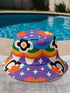 A reversible bucket hat with two distinct sides. Side one features a red and yellow zebra pattern with large pink and orange daisies. Side two has a deep purple background with white stars, rainbow scalloped bands in blue, green, and orange, and retro black and white flowers. The fabric has a visible textured linen weave. The hat is shown in a structured, professional studio shot against a white background, highlighting its 100% polyester durability.