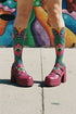 Crew-length socks featuring a vibrant 70s psychedelic pattern on a dark emerald green background. The design consists of vertical symmetrical medallions with hot pink scalloped edges, royal blue interior teardrops, and safety orange accents at the top ribbing. Fine white and light blue pointillist dots outline the major shapes. The heel and toe are solid black and reinforced. The fabric is a smooth. The pattern is maximalist and colorful, showcasing a retro aestheti.