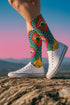 Crew-length socks featuring a vibrant 70s psychedelic pattern on a dark emerald green background. The design consists of vertical symmetrical medallions with hot pink scalloped edges, royal blue interior teardrops, and safety orange accents at the top ribbing. Fine white and light blue pointillist dots outline the major shapes. The heel and toe are solid black and reinforced. The fabric is a smooth. The pattern is maximalist and colorful, showcasing a retro aestheti.