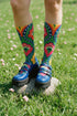 Crew-length socks featuring a vibrant 70s psychedelic pattern on a dark emerald green background. The design consists of vertical symmetrical medallions with hot pink scalloped edges, royal blue interior teardrops, and safety orange accents at the top ribbing. Fine white and light blue pointillist dots outline the major shapes. The heel and toe are solid black and reinforced. The fabric is a smooth. The pattern is maximalist and colorful, showcasing a retro aestheti.