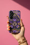 A matte finish phone case featuring a 2D optical illusion of a textured, purple crochet Ouija board planchette. The design includes intricate gold faux-embroidered borders, the words YES and NO, a central all-seeing eye, and a botanical leaf motif in the center circle. Lilac purple flowers and green leaves decorate the bottom half. The case has black rubber lining around the edges and camera cutouts. It looks like a thick, handmade yarn textile but is completely flat and smooth.